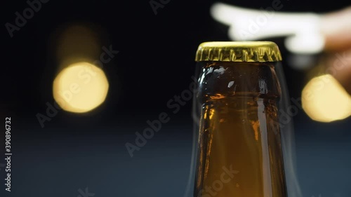 Close up male hand opening the bottle of beer on blue background with blurry lights. Foam rising and overflowing. The camera slowly moving on the slider