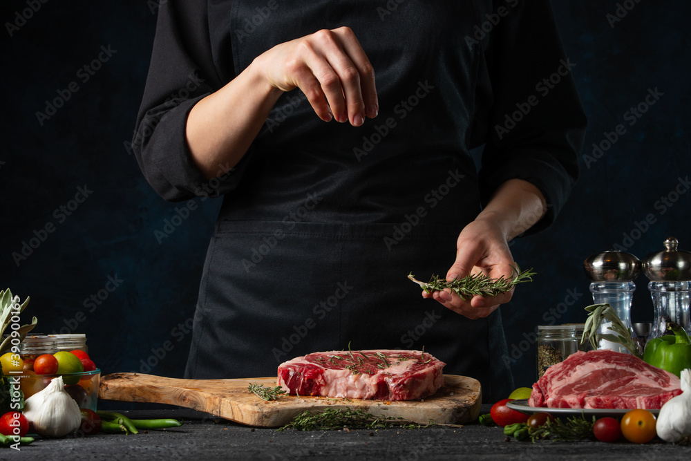 The chef in black uniform pours rosemary on raw meat steak on ingredients background. Frozen motion. Backstage of preparing restaurant delicious food with spices. Concept of cooking process.