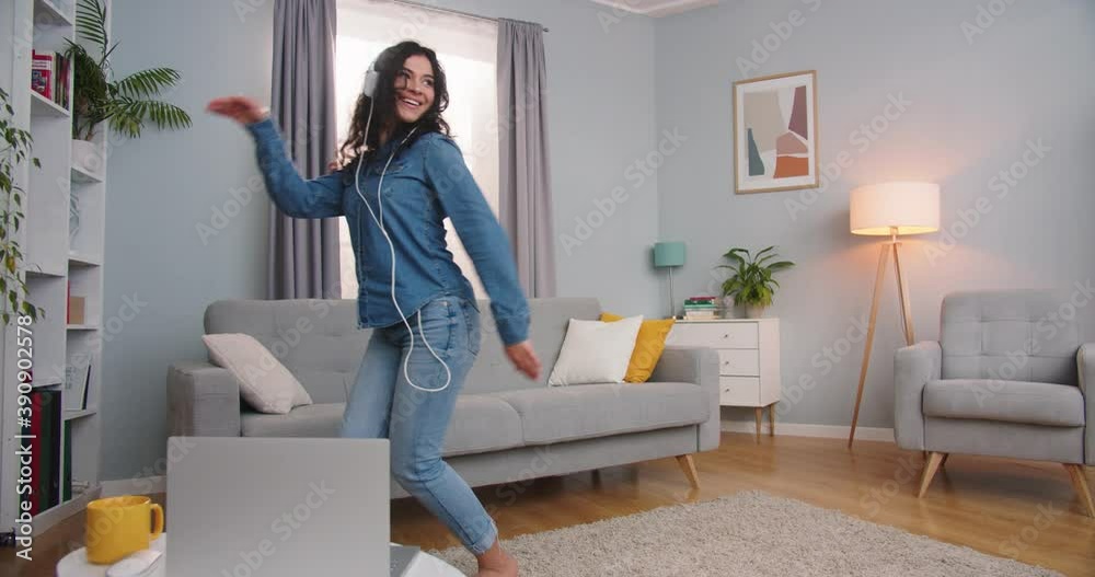 Cheerful Caucasian female in headphones enjoying song and dancing in living room. Pretty energetic young woman listening to music at home indoors. Entertainment, technology, leisure time. Cheerful Caucasian female in headphones enjoying song and dancing in living room. Pretty energetic young woman listening to music at home indoors. Entertainment, technology, leisure time.