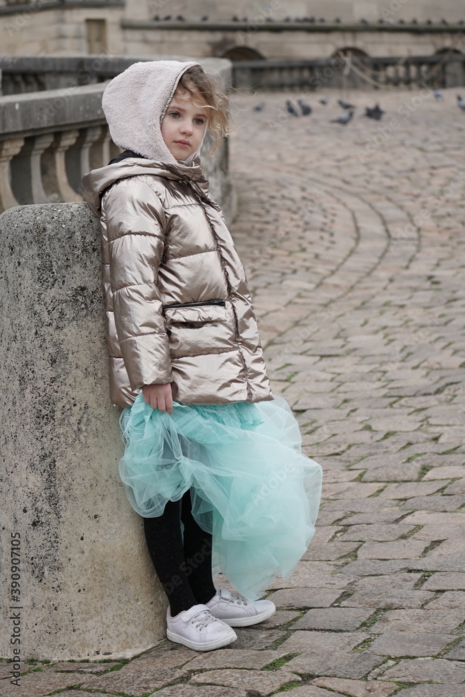The fashion girl wearing winter coat and beautiful dress standing against the old stone wall. Dreaming beauty in the french city The fashion girl wearing winter coat and beautiful dress standing against the old stone wall. Dreaming beauty in the french city