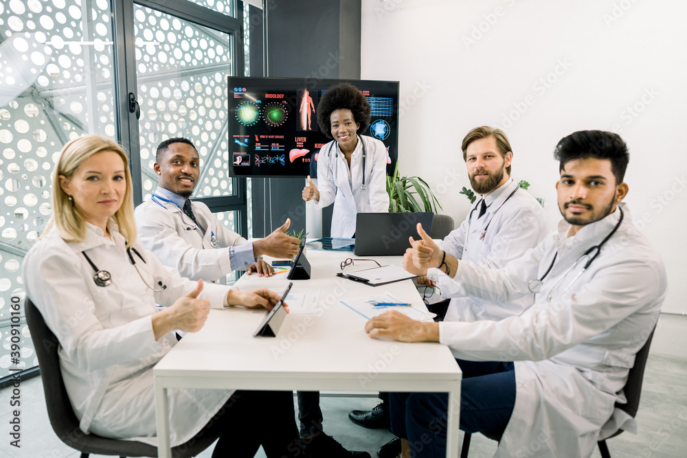 Fototapeta premium Team of smiling multiethnic doctors, Caucasian, African and Indian, having brainstorming session in the meeting room, sitting at the table and showing thumbs up to camera