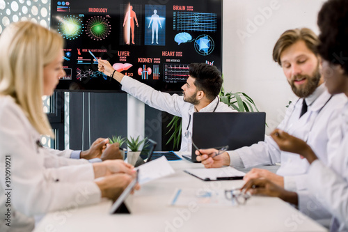 Young Indian doctor advises his multiethnical colleagues during a medical meeting, pointing on big digital screen with information and pictures about desease symptoms and spread