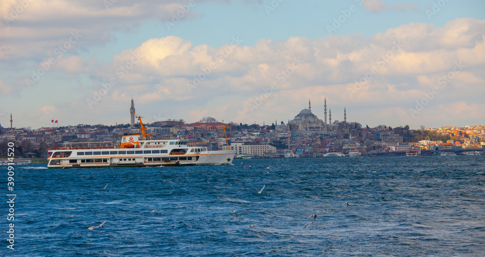 Classic retro ferry sailing towards Besiktas district in the European side of Istanbul on a cloudy day with Galata Tower in the background. Seagulls are flying around.