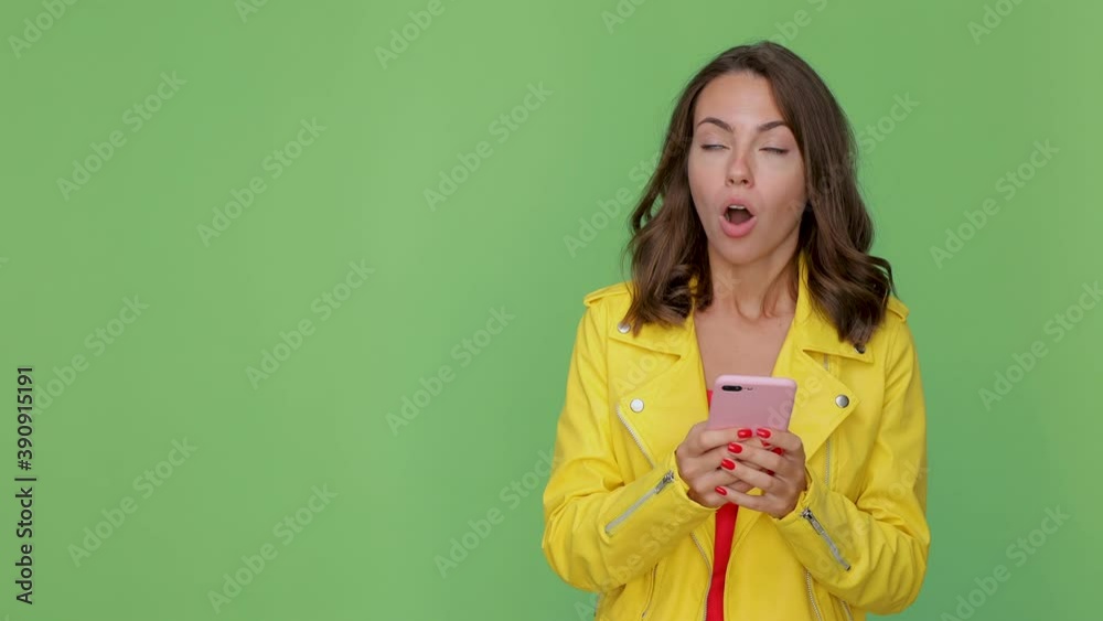 Shocked young woman 20s in yellow leather jacket posing isolated on green background studio. People lifestyle concept. Using mobile phone typing sms message point hand aside say wow showing thumb up Shocked young woman 20s in yellow leather jacket posing isolated on green background studio. People lifestyle concept. Using mobile phone typing sms message point hand aside say wow showing thumb up