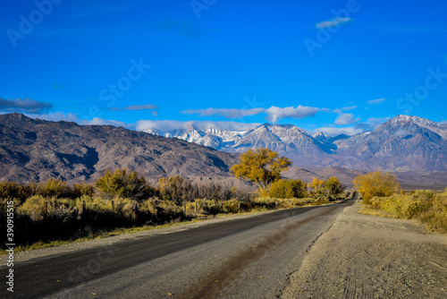 rural road to mountains with autumn trees and snow clouds in blue sky landscape