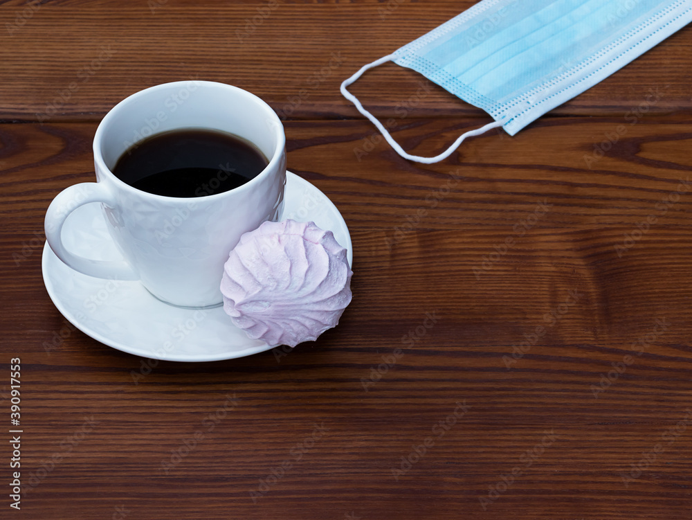 Obraz premium White cup of coffee and pink zefir on the wooden background. Traditional russian sweets like marshmallow. Copy space. Medical mask on the side.