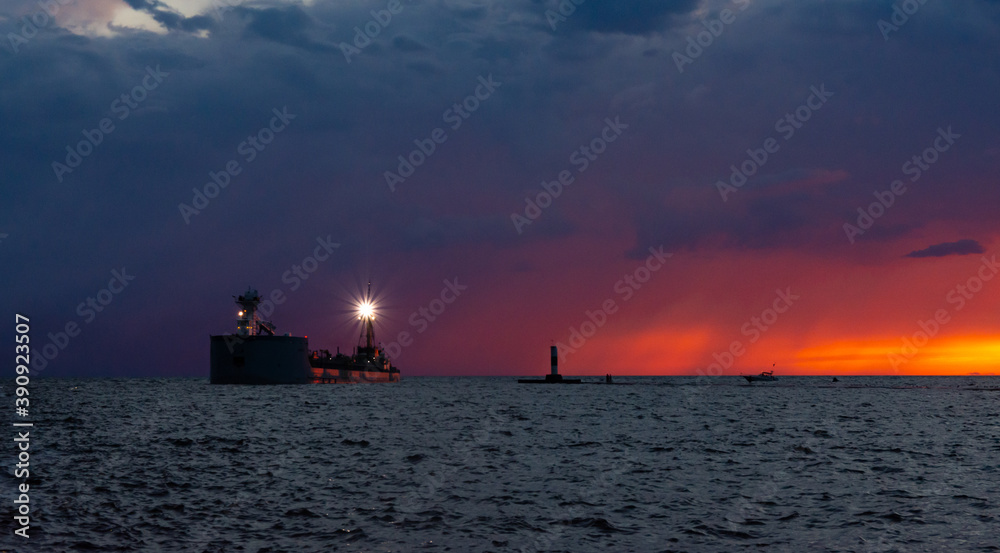 A dramatic sunset and rain storm over Lake Michigan provide a colorful backdrop to a barge sailing past the north pier at Grand Haven, Michigan.
