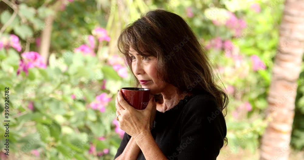 Attractive asian woman with long dark hair and wearing black top drinking coffee in medium shot on a lanai. Bright background, with tropical green and purple flowers. Attractive asian woman with long dark hair and wearing black top drinking coffee in medium shot on a lanai. Bright background, with tropical green and purple flowers.