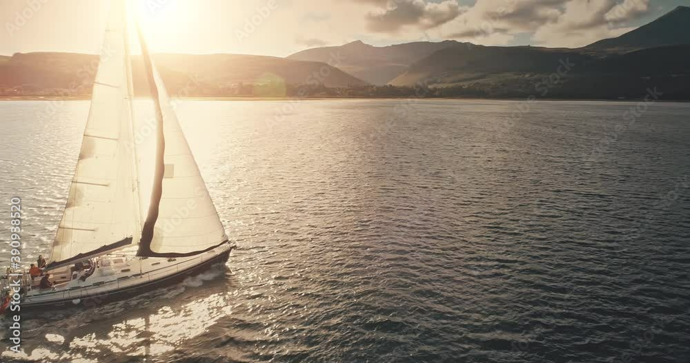 Sun reflection at sailboat on ocean bay aerial. Summer sea cruise on ship at green mountains island of Arran on yachts with passengers. Scotland sea coast water with vessel. Cinematic drone shot