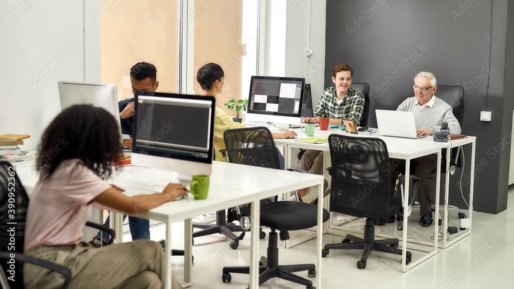 Fototapeta premium Full length shot of aged man, senior intern typing, using laptop while sitting at desk, working together with other young employees in modern office