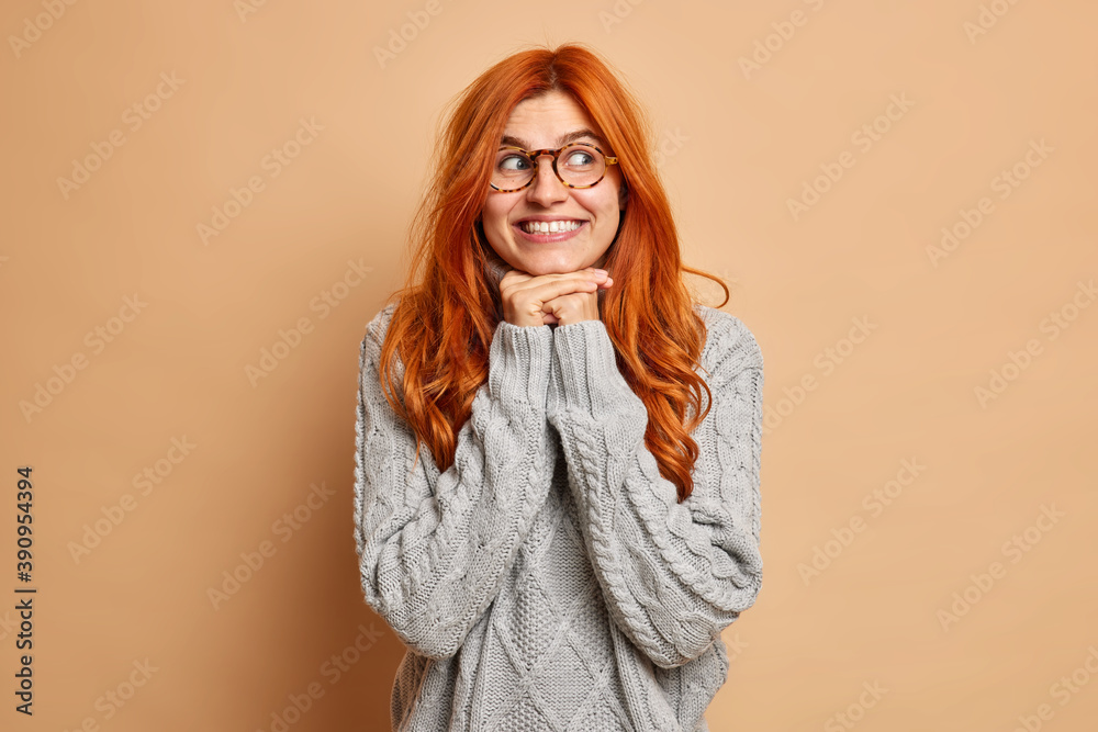 Good looking young woman with red hair keeps hands under chin smiles toothily concentrated aside wears spectacles and sweater isolated over brown background. Foxy lovely girl looks dreamily. Good looking young woman with red hair keeps hands under chin smiles toothily concentrated aside wears spectacles and sweater isolated over brown background. Foxy lovely girl looks dreamily.