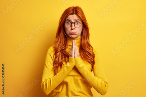Photo of desperate displeased woman with red hair keeps palms pressed together in pray gesture asks for apology looks with supplication dressed in casual turtleneck isolated over yellow background.
