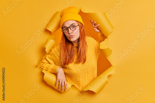 Lovely redhead young woman with round lips dressed in hat and sweater has flirting expression dressed in yellow clothing stands through torn paper background in hole. Facial expressions concept © wayhome.studio 