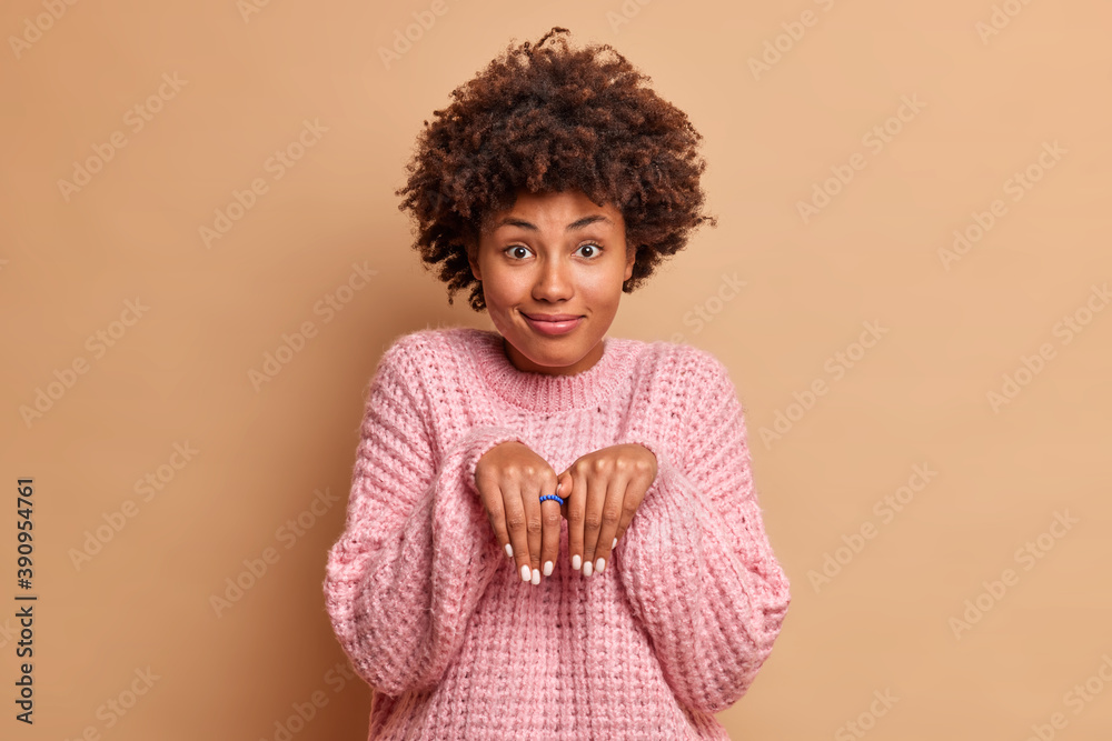 Funny dark skinned young woman makes rabbit paws and looks with innocent expression at camera plays with children has childish look dressed in knitted warm sweater isolated over brown background Funny dark skinned young woman makes rabbit paws and looks with innocent expression at camera plays with children has childish look dressed in knitted warm sweater isolated over brown background