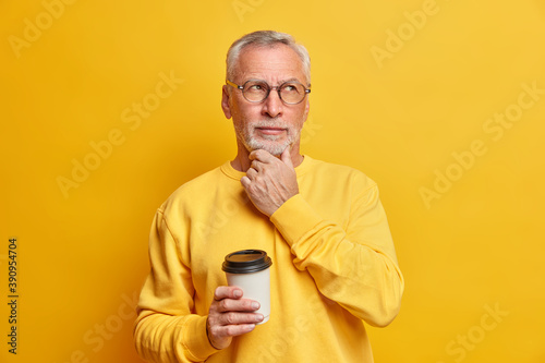 Pensive bearded male pensioner holds chin and concentrated aside drinks takeaway coffee and has break dressed in casual jumper isolated over yellow background. Thoughtful grandfather indoor. © wayhome.studio 