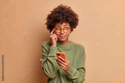 Pensive dark skinned young ethnic woman keeps index finger on temple tries to remember telephone number of someone holds modern smartphone and concentrated aside wears glasses casual jumper. © wayhome.studio 