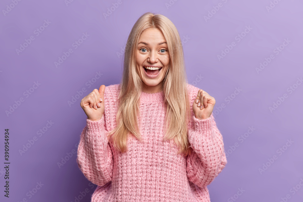 Positive blonde woman clenches fists and waits for something special happen laughs happily expresses good emotions dressed in loose knitted sweater isolated over purple background. Joy concept Positive blonde woman clenches fists and waits for something special happen laughs happily expresses good emotions dressed in loose knitted sweater isolated over purple background. Joy concept