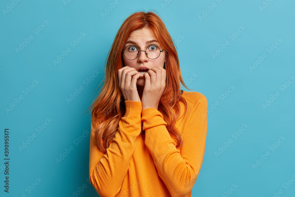 Puzzled surprised redhead Caucasian woman bites finger nails keeps mouth opened gasps from amazement dressed in casual jumper isolated over blue background. Human emotions and reaction concept Puzzled surprised redhead Caucasian woman bites finger nails keeps mouth opened gasps from amazement dressed in casual jumper isolated over blue background. Human emotions and reaction concept