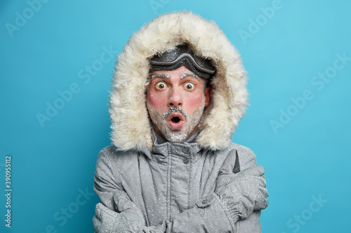 Cuadro en lienzo Stupefied frozen man with red face trembles from cold embraces himself to feel warm stares bugged eyes at camera wears snowboarding goggles isolated over blue studio background