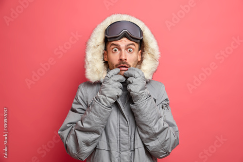 Winter hobby and recreation concept. Worried unshaven Caucasian man stares bugged eyes at camera dressed in outerwear wears snowboarding goggles finds out shocking news isolated over pink background.