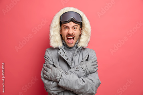 Emotional European man trembles from cold and shouts angrily keeps arms crossed dressed in winter jacket goes snowboarding during frosty day isolated over pink studio background. Mad angry skier