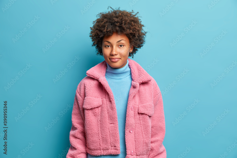 Fashionable dark skinned woman has curly hair dressed in pink coat looks happily gazes confident at camera isolated on blue background. Good looking Afro American teenage girl has pleasaed expression - obrazy, fototapety, plakaty 