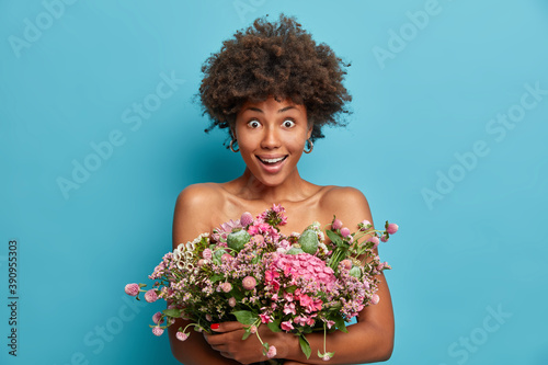 Greatly surprised dark skinned woman poses with bare shoulders holds big bunch of flowers gets bouquet from husband has pleased face expression isolated over blue background. Celebration concept
