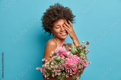 Photo of pleased dark skinned female model smiles gently covers face with hand hides naked body with big bouquet received from beloved person models against blue background. Happiness concept