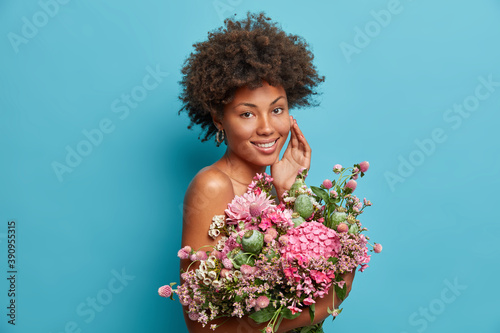 Adorable curly haired woman stands with bare shoulders touches face gently holds big beautiful bouquet of flowers expresses positive emotions isolated over blue background. Natural beauty concept