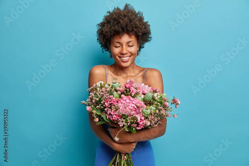 Pretty curly haired young woman glad to get proposal from boyfriend holds big bunch of flowers closes eyes with pleasure isolated over blue background. Happy Afro American lady embraces bouquet