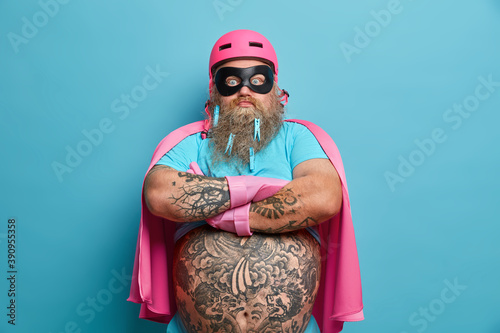 Photo of self assured bearded man keeps tattooed arms folded and stands in assertive pose with big fat belly wears helmet mask and cape pretends being super hero isolated over blue background. photo