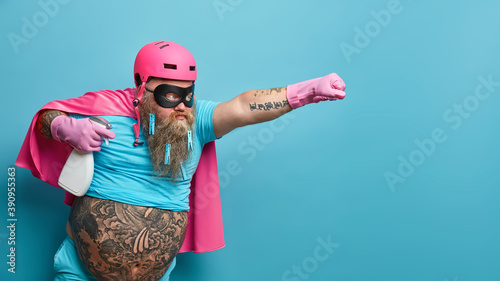 Horizontal shot of self confident male hero wears pink helmet cloak and rubber gloves holds bottle of detergent ready to help you with cleaning stretches arm as going to fly isolated over blue wall