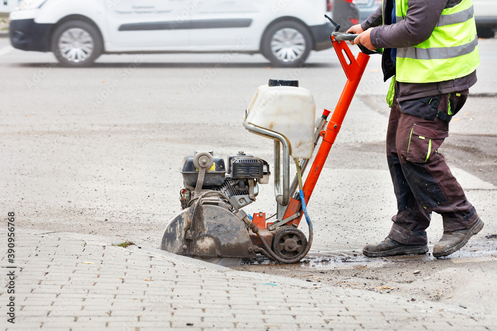 A worker cuts the old asphalt with a gas saw on the carriageway against the backdrop of a city street.