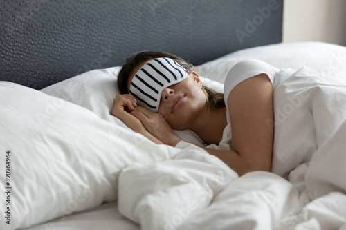 Stylish nightwear. Comfy sleeping mask helping young woman tourist traveler enjoy good healthy night sleep at new place in hotel suite room, millennial woman napping at wide king size bed in eye mask