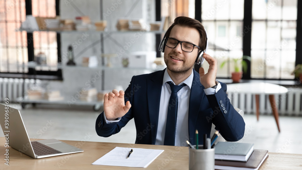 Relaxed young calm handsome smiling businessman manager in formal wear and eyeglasses sitting at workplace, enjoying stress free pause time listening favorite music tracks in modern earphones. Relaxed young calm handsome smiling businessman manager in formal wear and eyeglasses sitting at workplace, enjoying stress free pause time listening favorite music tracks in modern earphones.