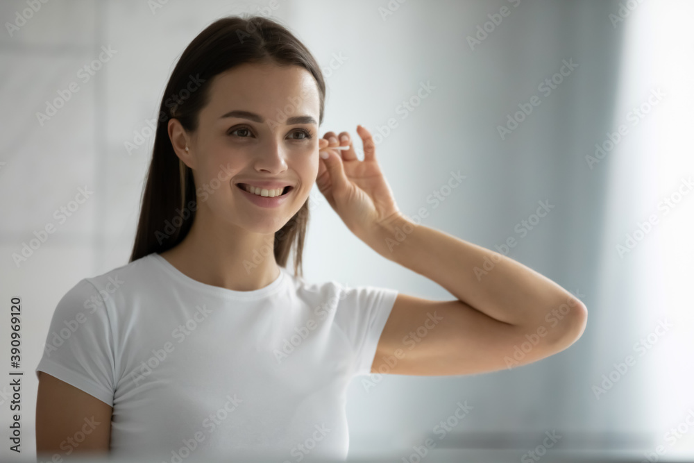 Gentle bodycare. Pleasant smiling young woman standing at bathroom near mirror concentrated on cleaning q-tipping her ears carefully using cotton swab bud, devoting time to everyday morning hygiene Gentle bodycare. Pleasant smiling young woman standing at bathroom near mirror concentrated on cleaning q-tipping her ears carefully using cotton swab bud, devoting time to everyday morning hygiene