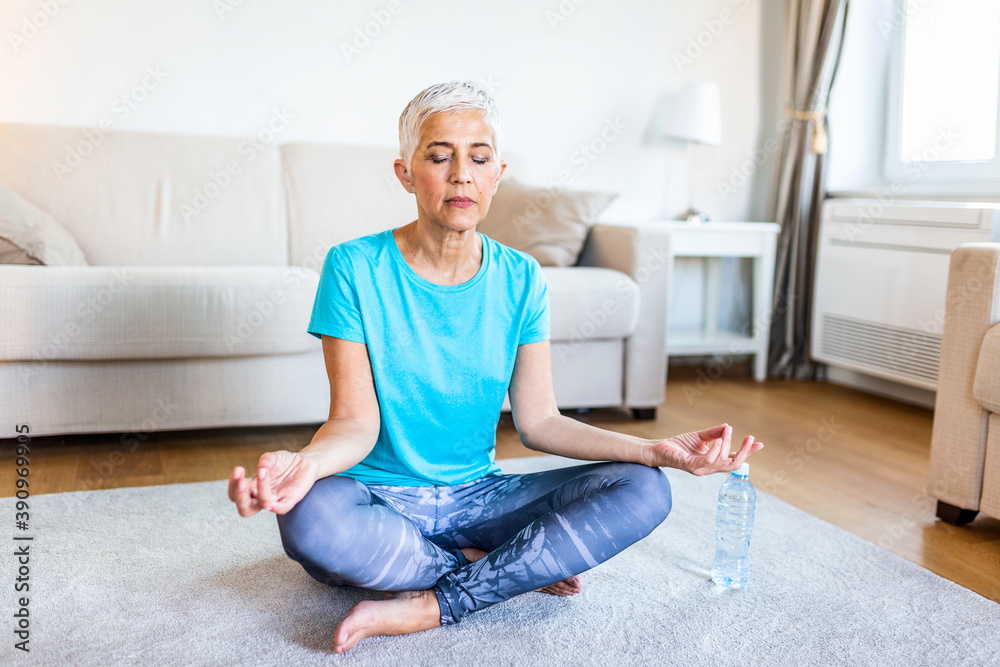Senior woman practicing yoga, sitting in Half Lotus exercise, Ardha Padmasana pose, working out, wearing sportswear, meditation session, indoor full length, home interior Senior woman practicing yoga, sitting in Half Lotus exercise, Ardha Padmasana pose, working out, wearing sportswear, meditation session, indoor full length, home interior