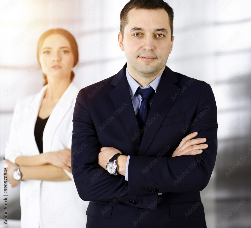 Portrait of a friendly middle aged businessman in a dark blue suit, standing with crossed arms together with a colleague in a sunny modern office. Modern business concept Portrait of a friendly middle aged businessman in a dark blue suit, standing with crossed arms together with a colleague in a sunny modern office. Modern business concept