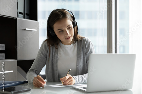 Work and study. Interested millennial woman employee trainee intern sitting at desk wearing wireless headphones watching webinar on computer screen writing up valuable information on paper sheet photo
