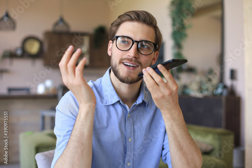 Confident business man in glasses holding smartphone near mouth for recording voice message or activating digital assistant.