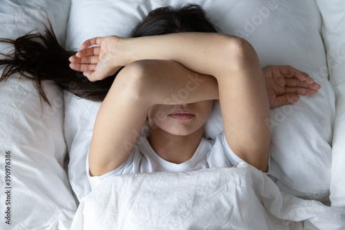 Leave me alone. Top view of capricious girl teenager or young female lying in cozy bed resisting unwilling to wake up early in the morning hiding face under crossed hands covering eyes from sunlight