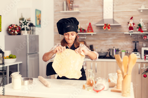 Child wearing apron and bonette on christmas days Cheerful happy cute girl while prepearing delicious cookies for christmas celebration in kitchen with christmas tree in the background. photo