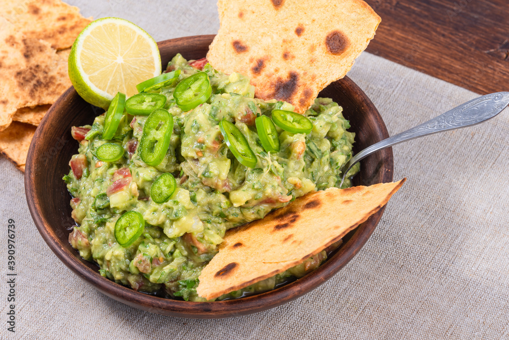 Homemade guacamole - a traditional Mexican appetizer of avacado, onion and tomato in a bowl with slices of toasted tortilla with avacado and lime on a rustic napkin on a wooden table Homemade guacamole - a traditional Mexican appetizer of avacado, onion and tomato in a bowl with slices of toasted tortilla with avacado and lime on a rustic napkin on a wooden table