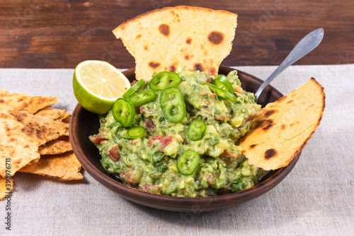 Homemade guacamole - a traditional Mexican appetizer of avacado, onion and tomato in a bowl with slices of toasted tortilla with avacado and lime on a rustic napkin on a wooden table