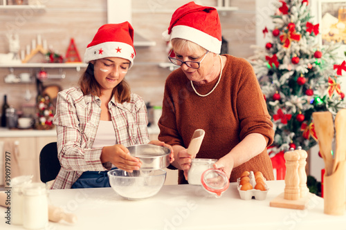 Multigeneration family doing cookies together on christmas day. Happy cheerful joyfull teenage girl helping senior woman preparing sweet buiscuits to celebrate winter holidays wearing santa hat.
