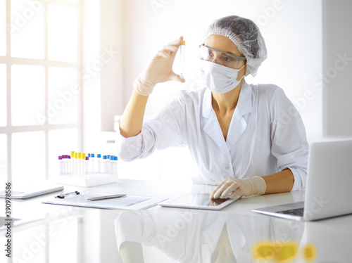 Professional female scientist in protective eyeglasses researching tube with reagents in sunny laboratory. Medicine and researching