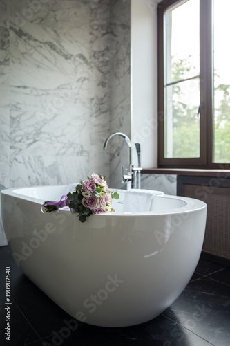 A delicate wedding bouquet of roses lies on the edge of a snow-white bathroom. Designer bathroom in the center of the bathroom. White artificial stone bathtub.