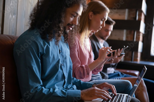 Gadget addiction. Diverse young people sitting on couch in public place involved in using different electronic devices, looking at screens, communicating online, avoiding real life talks conversations