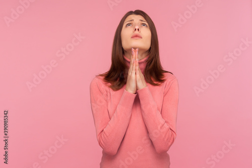 Lonely upset brunette woman looking up holding hands together, praying God, asking for health and support pleading for healing and forgiveness. Indoor studio shot isolated on pink background