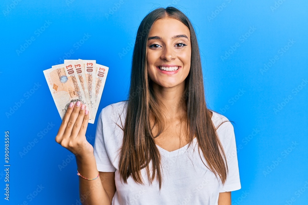 Young hispanic woman holding united kingdom pounds looking positive and happy standing and smiling with a confident smile showing teeth Young hispanic woman holding united kingdom pounds looking positive and happy standing and smiling with a confident smile showing teeth